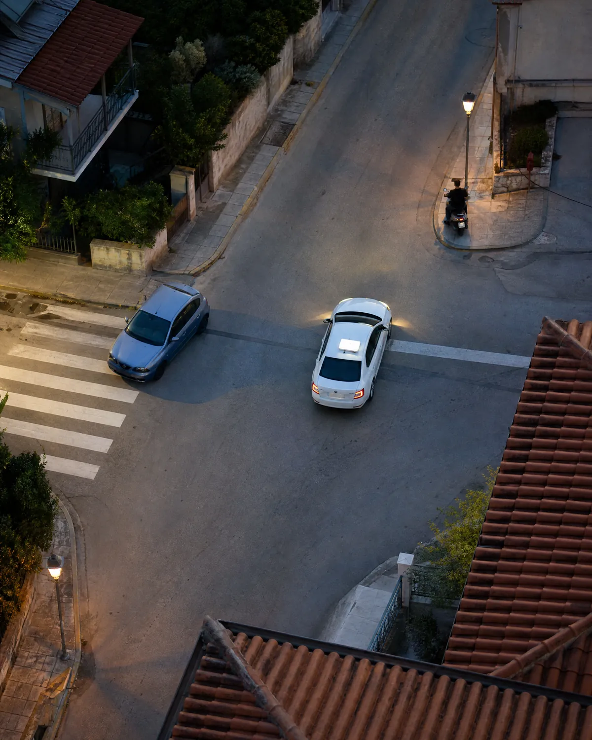 Σταυροδρόμι επαρχιακής πόλης από ψηλά νωρίς το πρωί: αυτοκίνητο σχολής οδηγών λίγο πέρα από τη γραμμή του στοπ, μικρό αυτοκίνητο παρκαρισμένο πάνω στη διάβαση πεζών και οδηγός σκούτερ χωρίς κράνος στη γωνία, με κεραμοσκεπές γύρω.