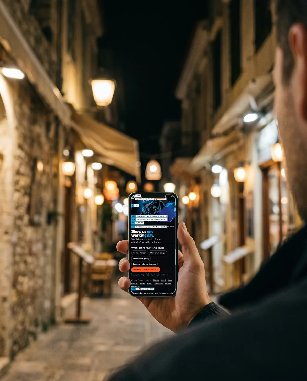 A hand holding a phone at night in a narrow old-town street; on screen, the netex.gr home page with its X-ray labels.